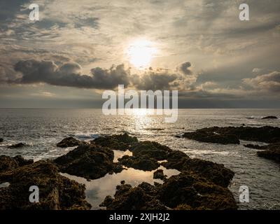 Sun shining through clouds over the ocean creating tide pools on the rocky coast Stock Photo