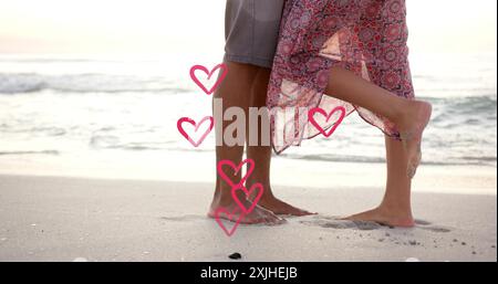 Image of hearts moving over legs of diverse couple in love on beach in summer Stock Photo