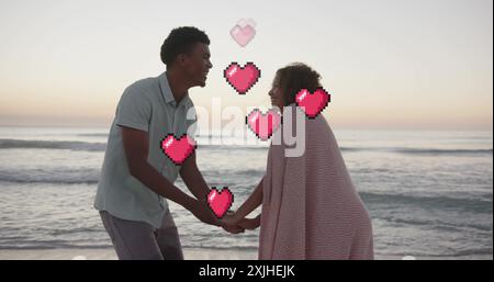 Image of hearts moving over diverse couple in love holding hands on beach in summer Stock Photo