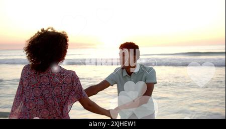 Image of hearts moving over diverse couple in love on beach in summer. Love, romance, valentine's day and vacation concept digitally generated image. Stock Photo