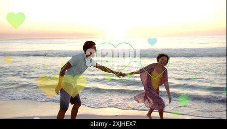 Image of hearts moving over diverse couple in love dancing on beach in summer Stock Photo