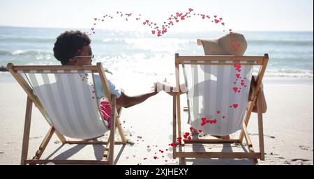 Image of hearts moving over diverse couple in love in deckchairs on beach in summer Stock Photo