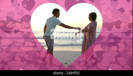 Image of hearts moving over diverse couple in love dancing on beach in summer Stock Photo