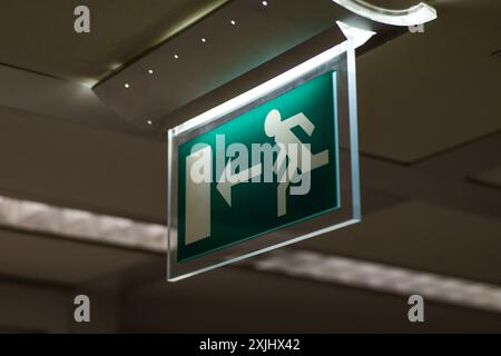 The image captures an illuminated exit sign mounted on the ceiling. The sign features a green background with a white figure running towards a door, a Stock Photo