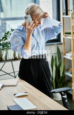 A middle aged businesswoman with short hair, during menopause, stands in an office engaged in negotiations on a cell phone. Stock Photo