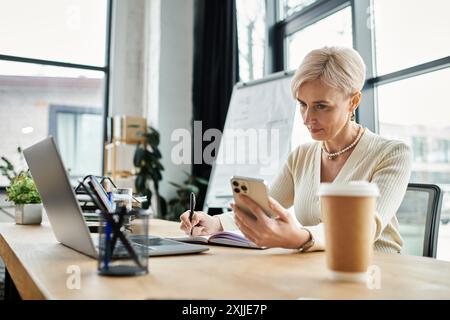 A middle-aged businesswoman with short hair is seated at a table using a laptop and a cell phone in a modern office setting. Stock Photo