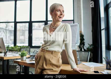 A middle-aged businesswoman with short hair confidently stands on a desk in a modern office, immersed in her work. Stock Photo