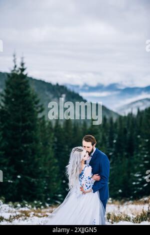 groom in a blue suit and bride in white, embroidered with blue pattern, dress on a background of green pine forests in the mountains of the Carpathian Stock Photo