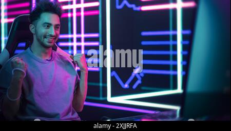 Excited Handsome Man Playing Video Games with Friends on a Computer, Surrounded by Cyberpunk Style Background. Young Gamer Winning the Round and Celebrating Stock Photo