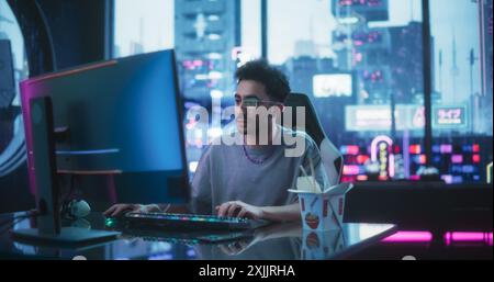 Handsome Young Man Eating Take-Away Asian Food, while Working on a Desktop Computer in a Cyberpunk Neon Room. Futuristic Hacker Writing Software Code and Wearing Innovative Augmented Reality Glasses Stock Photo
