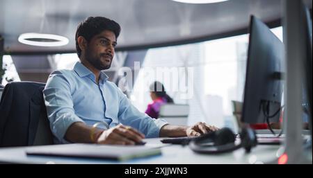 Portrait of a South Asian Financial Securities Trader Working on Desktop Computer in a Modern Office. Handsome Indian Accountant Bookkeeping Financial Affairs of a Business Venture Stock Photo