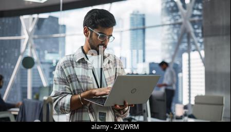 Indian Male Business Analyst Standing with Laptop Computer in a Modern Office. Successful Man Assessing Technological Risks And Developing Strategies For Company Growth and Expansion Stock Photo