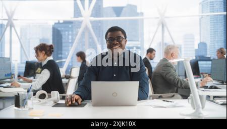 Succesful Black Man Typing On Laptop Computer In Bright Scyscraper Office With Diverse Employees In Downtown. Cheerful Male Programmer Looking At Camera And Smiling In Fintech Startup Workspace. Stock Photo