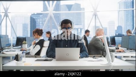 Successful Black Man Wearing Headphones And Starts Typing On Laptop Computer In Skyscraper Office In Downtown. Professional Male Software Developer Working On a New Project In Diverse Tech Startup. Stock Photo