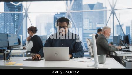 Successful Black Man Wearing Headphones And Starts Typing On Laptop Computer In Skyscraper Office In Downtown. Professional Male Product Manager Working On a New Project In Diverse Workspace. Stock Photo