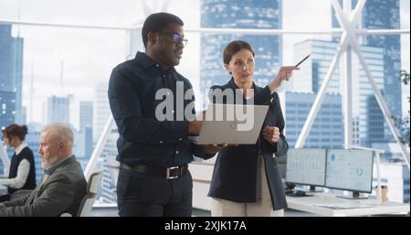 Businesspeople in Modern Office: Business Meeting of Two Managers Using Laptop. Female CEO and African American Operations Director Talk, Discuss Strategy, Implementing Marketing and Financial Plans. Stock Photo