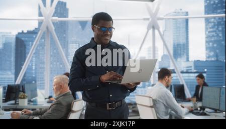 Handsome African American CEO Walking In Modern Skyscraper Office With Diverse Employees In Downtown. Succeful Black Man Using Laptop Computer To Go Through Tasks And Business Meetings In Tech Startup Stock Photo