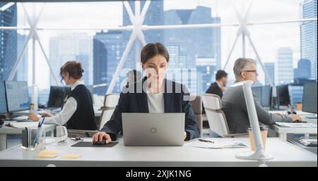 Professional Female Business Development Manager Typing On Laptop Computer In Bright Skyscraper Office In Downtown. Successful Caucasian Woman Working With Annual Report In Multiethnic Workspace. Stock Photo