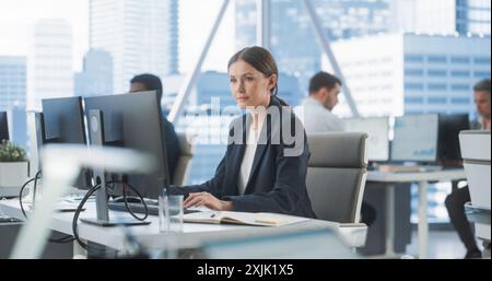 Focused Female Operations Specialist Typing On Desktop Computer In Diverse Corporate Office With Megapolis Window View. Professional Caucasian Woman Writing Emails to Clients, Scheduling Meetings. Stock Photo