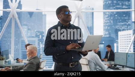 African American Financial Analyst Walking In Skyscraper Office With Diverse Employees In Downtown. Succeful Black Man Using Laptop Computer To Go Through Tasks And Business Mettings In Hedge Fund. Stock Photo