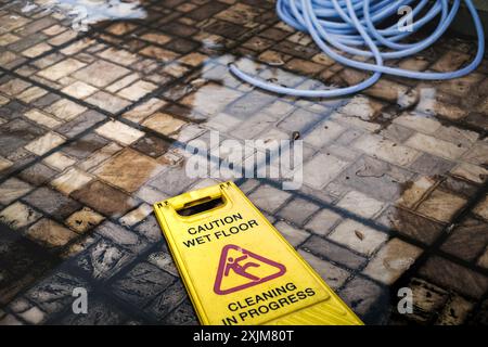 Yellow Caution Wet Floor Cleaning in Progress Sign and Gardening Hose Fell on Dirty Mossy Wet and Flooded Outdoor Floor Stock Photo