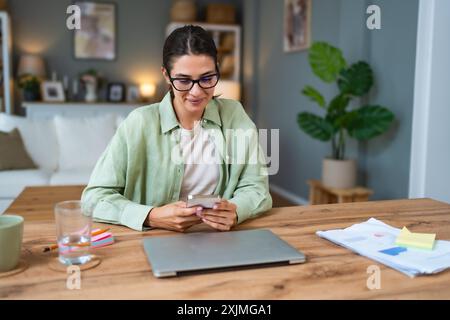 Young business woman using break from working in home office for surfing on social media. Female freelance designer professional using smartphone for Stock Photo