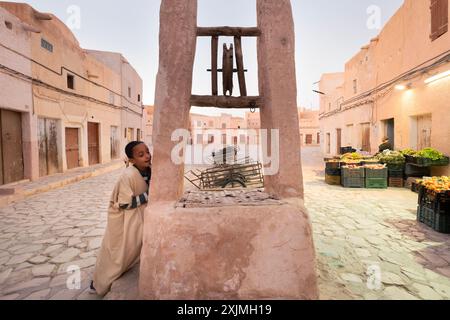 Young boy is standing by an old well in a traditional north african market, with a vendor selling fruits and vegetables in the background, Algeria Stock Photo