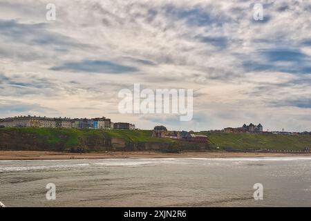 A coastal town with buildings on a cliff overlooking a sandy beach and the ocean under a cloudy sky. Stock Photo