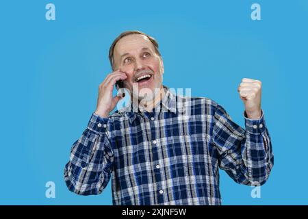 Portrait of an extremely happy aged senior man is talking on phone and celebrating success. Isolated on blue. Stock Photo