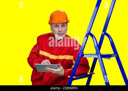 Senior male worker holding tablet pc leans on a blue ladder. Isolated on yellow. Stock Photo