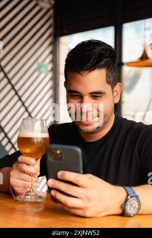 A smiling man in a black shirt enjoys a glass of beer while using his smartphone in a modern bar. The environment has contemporary decor, and the man Stock Photo