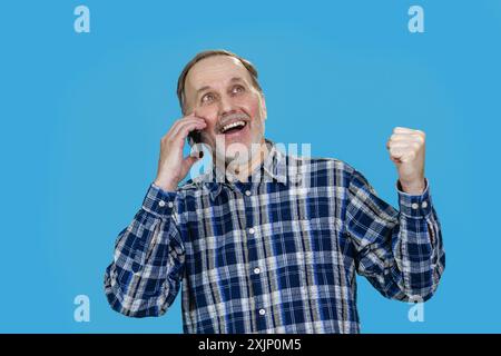 Portrait of an extremely happy aged senior man is talking on phone and celebrating success. Isolated on blue. Stock Photo