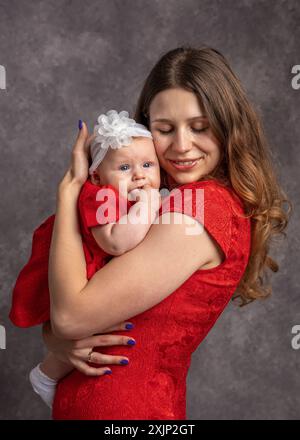 Mom gently hugs her little daughter. Dressed in red dresses. Gray background.Studio shooting. Mother's love. Newborn care. Family concept Stock Photo