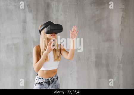 Blonde woman in white tank top and jeans with VR headset in front of gray wall, interacting with virtual world. Hands extended, immersed in VR. Stock Photo