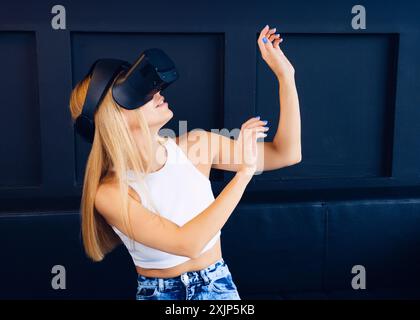 A woman wearing a virtual reality headset stands in front of a dark blue wall, reaching out with her hands as if interacting with something in the Stock Photo