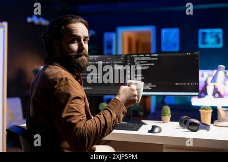 Portrait of happy computer technician enjoying hot tea while implementing security measures to protect company systems and data. Joyful IT worker holding coffee mug and debugging code Stock Photo
