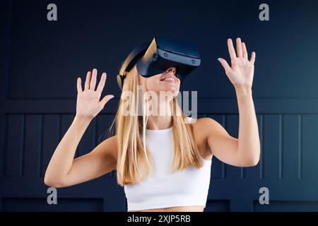 Woman wearing a virtual reality headset looks upwards with her arms outstretched over black wall in gaming room. Technologies and entertainment  Stock Photo