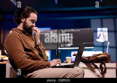 IT specialist working from home using laptop, testing and deploying programs and systems. Programmer checking code displayed on computer monitors, sitting at desk chair in apartment Stock Photo