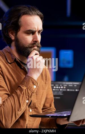 Pensive cybersecurity expert looking confused while using laptop to fix company security vulnerabilities. Contemplative developer feeling worried about potential cyberattacks, close up shot Stock Photo
