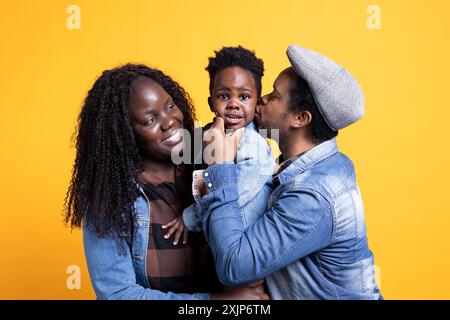 Affectionate father kissing his little son on the cheek, happy loving family of three posing in studio. African american couple holding their toddler and bonding together, showing love and respect. Stock Photo