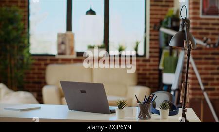 Cozy stylish empty apartment lit by sunlight coming from big windows, TV left open in background and laptop on table desk. Bright modern living room with brick walls and hygge eclectic interior decor Stock Photo