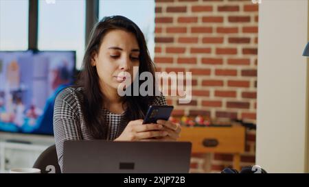 Smiling person checking mobile phone notifications at computer desk, taking break from work. Indian woman in cozy apartment pausing job tasks to read messages on cellphone, camera B close up Stock Photo