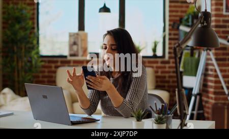 Teleworker checking her phone notifications at home in front of laptop, taking break from work. Woman in cozy apartment office taking pause from job tasks to read messages on smartphone, camera B Stock Photo