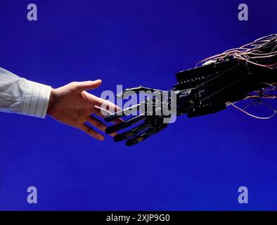 A human hand, wearing a white shirt with thin stripes, reaches out towards a mechanical robotic arm. The arm is black and made of metal and wires. The Stock Photo