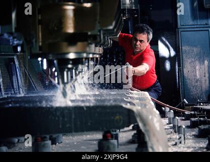 A worker wearing a red shirt operates a CNC machine, carefully monitoring the process as it cuts and shapes a large piece of metal. The machine is spr Stock Photo
