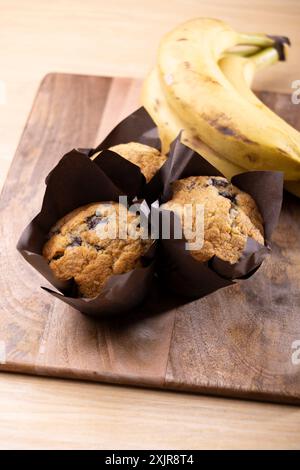 Freshly baked banana chocolate chip muffins wrapped in brown parchment paper, placed on a wooden cutting board alongside a bunch of ripe bananas. Stock Photo