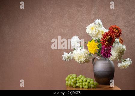 A large bouquet of dahlias in a gray clay vase on a wooden table. Nearby lies a bunch of green grapes. Orange background. White, yellow, red flowers. Stock Photo