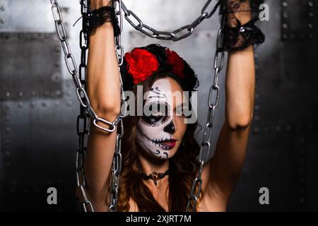 Woman wearing intricate sugar skull makeup and a floral headpiece holds up chains in a dark, industrial setting.  Stock Photo