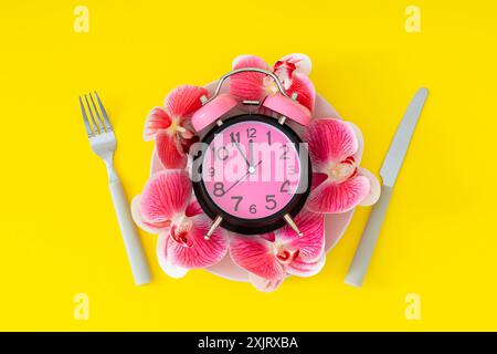Creative layout made with retro alarm clock, orchid flowers, pink plate, fork and knife on yellow background. Minimal time concept. Flat lay. Stock Photo