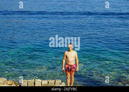 A young man who came out after swimming in the sea stands on the shore against the backdrop of clear water of a beautiful bright color (Montenegro) Stock Photo